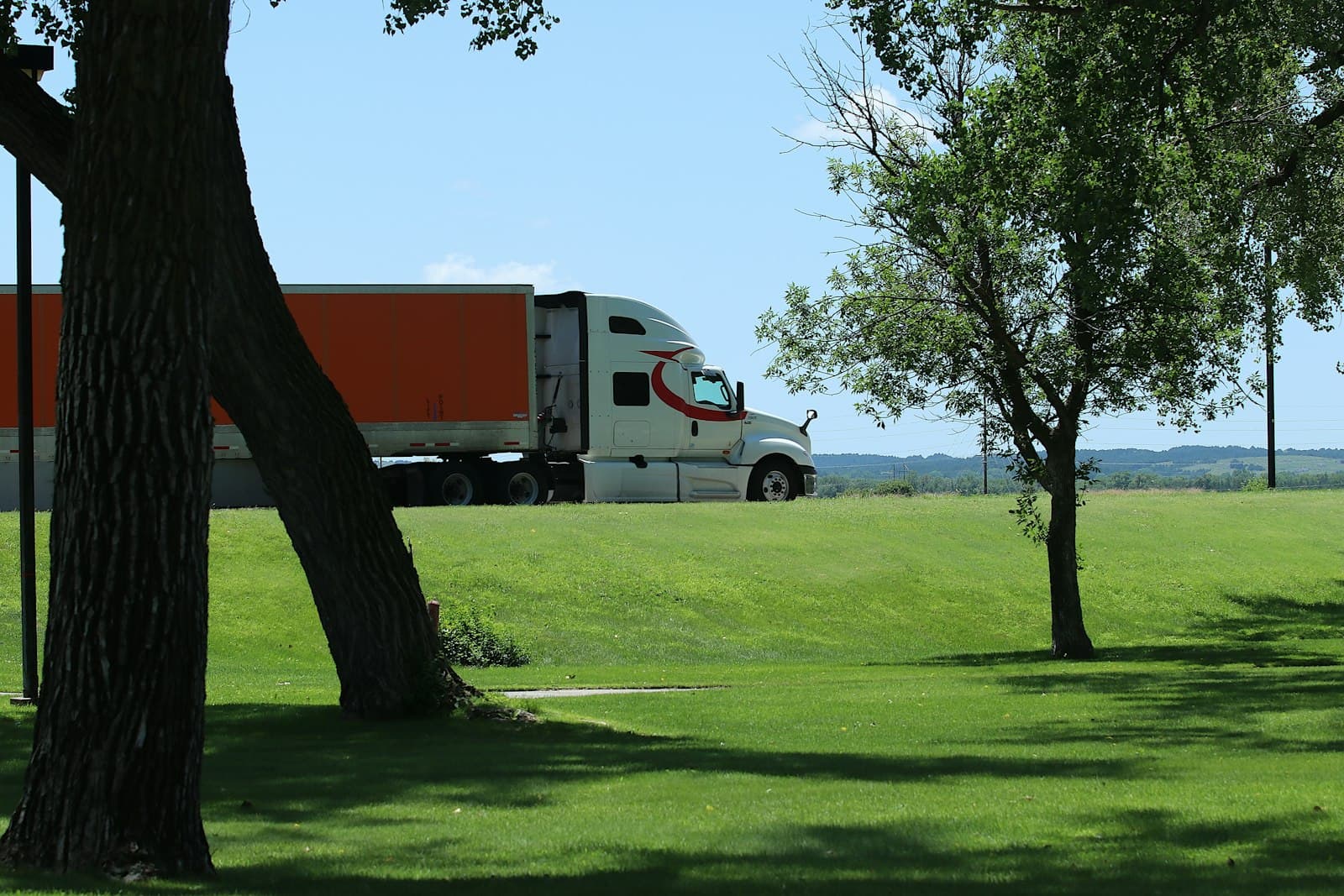 A white semi-truck with an orange trailer passing a tree-lined rest stop